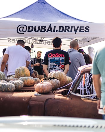 A group of people gather around an old rusty car that is decorated with numerous pumpkins. Above them is a canopy tent with the text '@DUBAI.DRIVES'. The setting seems lively, likely at an outdoor event or car show. Various individuals are engaged in conversation, with one person wearing a shirt featuring car graphics.