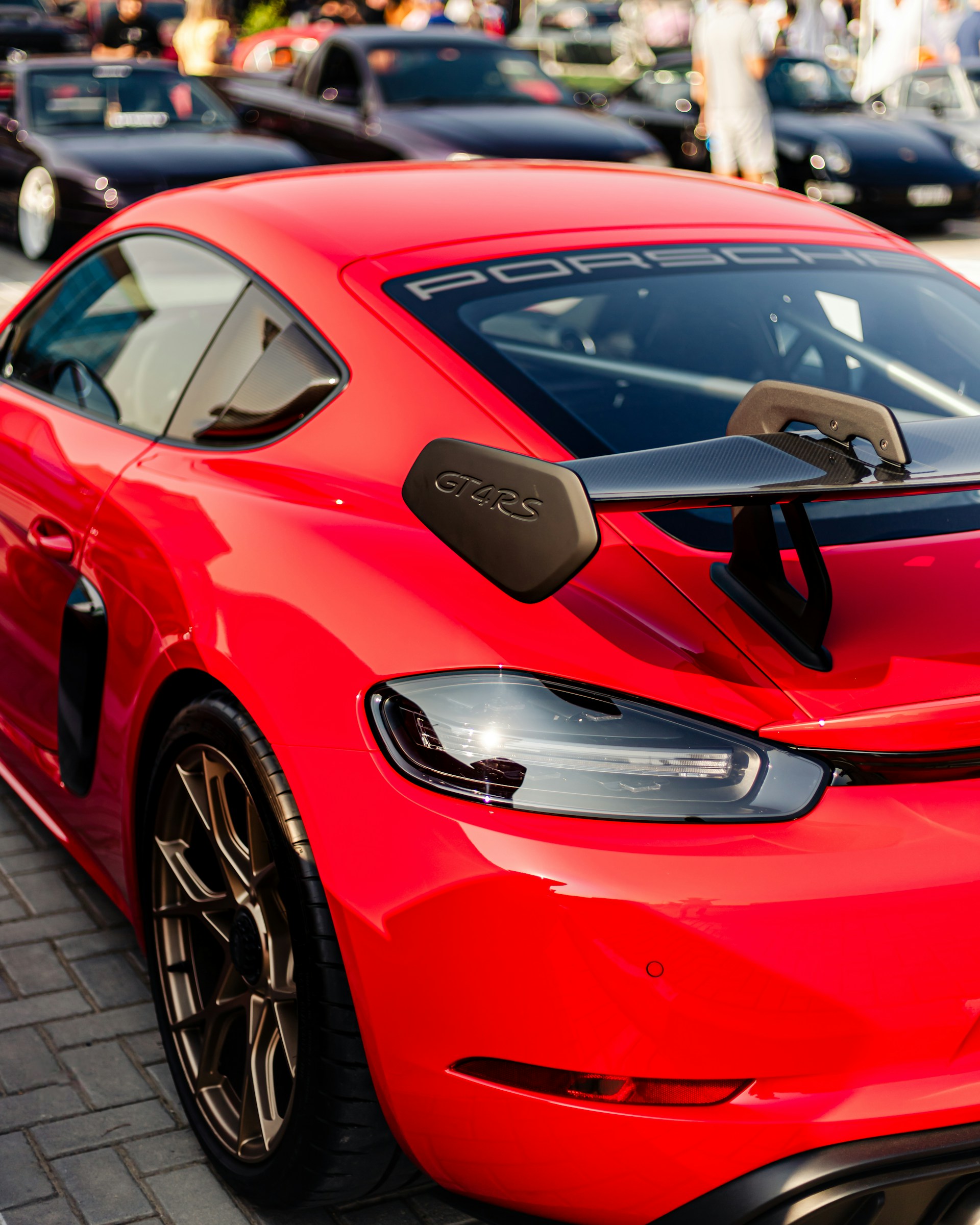 A bright red sports car is prominently displayed with a sleek design and a GT4 RS badge on its rear. The car is parked on a paved area, with several other cars visible in the background, including black vehicles with tinted windows. The crowd of people in the background suggests a social or automotive event.