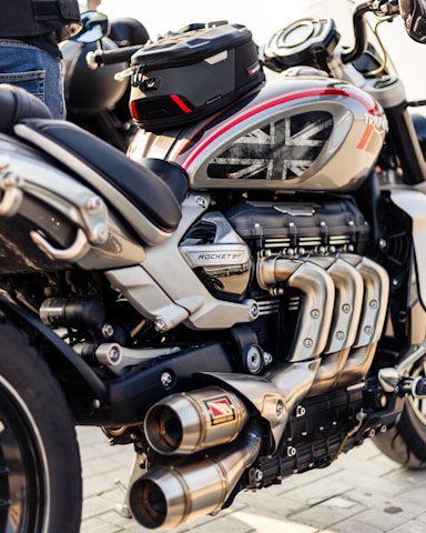 A close-up view of a powerful motorcycle featuring a sleek design with metallic and black components. The engine and exhaust system are prominently displayed, and a small bag is attached on top. The motorcycle tank displays a Union Jack design.