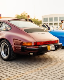 A classic burgundy Porsche car is parked on a paved area, with visible details including the rear lights, distinctive logo, and Dubai license plate. The scene appears to be at a car show or gathering, with a blue car partly visible next to it and people in casual clothing in the background.