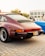 A classic burgundy Porsche car is parked on a paved area, with visible details including the rear lights, distinctive logo, and Dubai license plate. The scene appears to be at a car show or gathering, with a blue car partly visible next to it and people in casual clothing in the background.