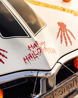 Decorated car hood with 'Happy Halloween' written in red, stylized as if painted with blood. There are also red handprint designs on the white hood, and a part of a pumpkin is visible. A Halloween-themed tape with skulls runs across the top.