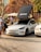 A white Tesla car is parked on a cobblestone surface with a rooftop tent labeled 'CHICARI Get Lost In Nature'. Surrounding the car is a person in a white shirt standing nearby, another person wearing a floral shirt and hat, and other cars parked nearby. The backdrop features a colorful, abstract mural with various shapes, and there is a white umbrella providing shade.