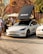 A white Tesla car is parked on a cobblestone surface with a rooftop tent labeled 'CHICARI Get Lost In Nature'. Surrounding the car is a person in a white shirt standing nearby, another person wearing a floral shirt and hat, and other cars parked nearby. The backdrop features a colorful, abstract mural with various shapes, and there is a white umbrella providing shade.