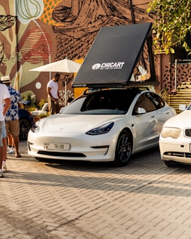 A white Tesla car is parked on a cobblestone surface with a rooftop tent labeled 'CHICARI Get Lost In Nature'. Surrounding the car is a person in a white shirt standing nearby, another person wearing a floral shirt and hat, and other cars parked nearby. The backdrop features a colorful, abstract mural with various shapes, and there is a white umbrella providing shade.