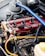 A detailed view of a car engine featuring a bright red valve cover with black ignition coils connected by wires. There is a prominent blue strut brace across the top and visible hoses and metal components surrounding the engine. The Ford branding is clearly visible.
