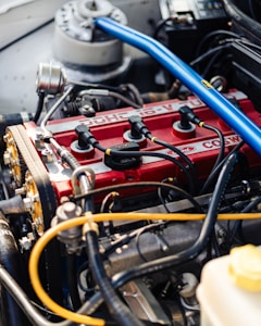A detailed view of a car engine featuring a bright red valve cover with black ignition coils connected by wires. There is a prominent blue strut brace across the top and visible hoses and metal components surrounding the engine. The Ford branding is clearly visible.