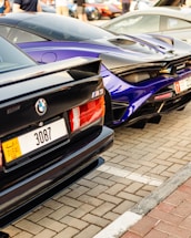 Two luxury cars, a black BMW M3 and a blue McLaren, are parked side by side on a paved parking lot. Both vehicles feature sleek, sporty designs, and the image captures their rear ends. The number plates are visible, showing a Dubai registration.