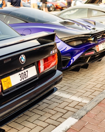 Two luxury cars, a black BMW M3 and a blue McLaren, are parked side by side on a paved parking lot. Both vehicles feature sleek, sporty designs, and the image captures their rear ends. The number plates are visible, showing a Dubai registration.