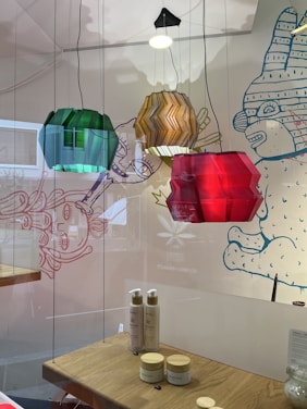 Hanging colored lamps in green, amber, and red are positioned in front of a decorative wall with whimsical cartoon-style drawings. Below the lamps, a wooden shelf displays skincare products, including bottles and jars, with a clean and minimalist setup.