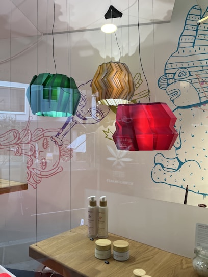Hanging colored lamps in green, amber, and red are positioned in front of a decorative wall with whimsical cartoon-style drawings. Below the lamps, a wooden shelf displays skincare products, including bottles and jars, with a clean and minimalist setup.