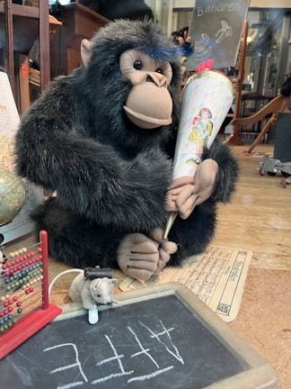 A plush gorilla is holding a decorative cone with a vintage illustration of a child. In front of it, an abacus and a small chalkboard with the word 'Affe' written are visible, along with a plush mouse holding a bottle. A globe and some papers are partially visible in the background.