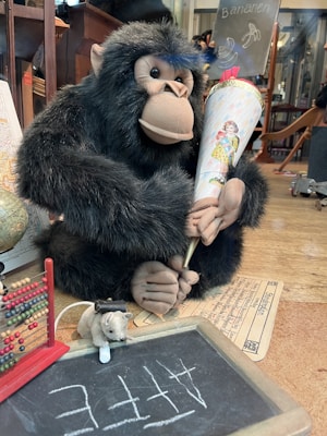 A plush gorilla is holding a decorative cone with a vintage illustration of a child. In front of it, an abacus and a small chalkboard with the word 'Affe' written are visible, along with a plush mouse holding a bottle. A globe and some papers are partially visible in the background.