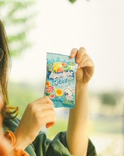 A person is holding up a package of Alpenliebe Jelly candy in a light blue wrapper. The package features colorful fruit illustrations and a cartoon character, creating a playful and inviting design.