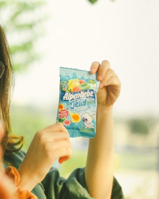 A person is holding up a package of Alpenliebe Jelly candy in a light blue wrapper. The package features colorful fruit illustrations and a cartoon character, creating a playful and inviting design.