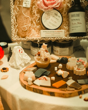 An assortment of handcrafted soaps and bath products arranged on a table. The display includes a cupcake-shaped soap, various small soaps shaped like animals and objects, and bath products in bottles and containers. A decorative setup with a mix of colors and textures is visible, highlighted by a large pink flower and packaging materials.