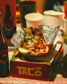 A serving of tacos with various toppings such as diced onions and cilantro, wrapped in aluminum foil, is placed on a table. Surrounding the tacos are beverage bottles and disposable cups. The packaging is colorful with a prominent label reading 'Taco'.