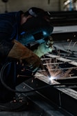 A person wearing a welding helmet and gloves is actively welding metal pieces, creating bright sparks. The scene is industrial, with dark tones dominating the environment.
