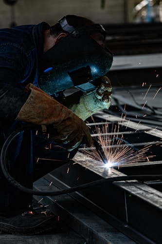A person wearing a welding helmet and gloves is actively welding metal pieces, creating bright sparks. The scene is industrial, with dark tones dominating the environment.