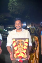 A man is holding a framed religious image adorned with a garland of red and yellow flowers. He appears to be participating in a ceremonial or festive event, with several people in traditional attire visible in the background. The event is taking place outdoors during the night.
