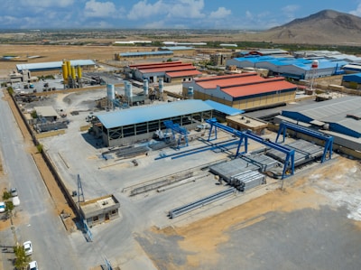 An industrial complex with multiple large buildings and structures, including warehouses with colorful roofs and various machinery, is situated in a dry, expansive landscape. The area is surrounded by roads and open spaces, with a distant view of a hilly terrain under a partly cloudy sky.