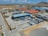 An industrial complex with multiple large buildings and structures, including warehouses with colorful roofs and various machinery, is situated in a dry, expansive landscape. The area is surrounded by roads and open spaces, with a distant view of a hilly terrain under a partly cloudy sky.