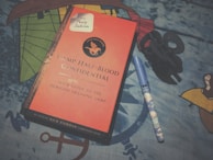 A book titled 'Camp Half-Blood Confidential' with a red cover and a circular emblem featuring a winged horse. Next to the book is a blue pen with a cartoon design and a black hair clip. The background is a patterned blue fabric.