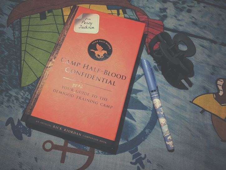 A book titled 'Camp Half-Blood Confidential' with a red cover and a circular emblem featuring a winged horse. Next to the book is a blue pen with a cartoon design and a black hair clip. The background is a patterned blue fabric.