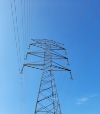 A tall, metal electricity transmission tower stretches upwards against a clear blue sky. Several power lines extend from the tower in multiple directions, emphasizing the structure's height and industrial design.