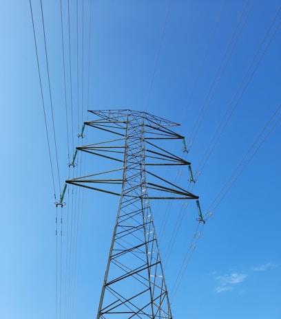A tall, metal electricity transmission tower stretches upwards against a clear blue sky. Several power lines extend from the tower in multiple directions, emphasizing the structure's height and industrial design.