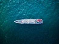 An aerial view of a sleek, modern ship navigating through deep blue-green ocean waters. The ship's deck features various equipment, vehicles, and structures arranged symmetrically.