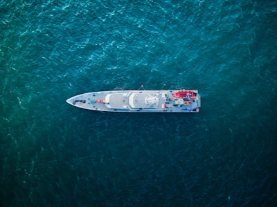 An aerial view of a sleek, modern ship navigating through deep blue-green ocean waters. The ship's deck features various equipment, vehicles, and structures arranged symmetrically.