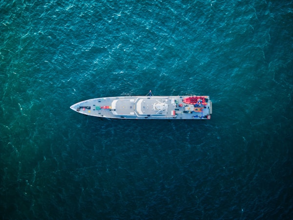 An aerial view of a sleek, modern ship navigating through deep blue-green ocean waters. The ship's deck features various equipment, vehicles, and structures arranged symmetrically.