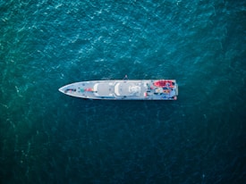An aerial view of a sleek, modern ship navigating through deep blue-green ocean waters. The ship's deck features various equipment, vehicles, and structures arranged symmetrically.