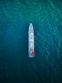 An overhead view of a large ship navigating through a vast expanse of deep blue ocean. The ship is centrally positioned, showcasing its streamlined design and various components visible on deck, including lifeboats and equipment.