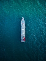 An overhead view of a large ship navigating through a vast expanse of deep blue ocean. The ship is centrally positioned, showcasing its streamlined design and various components visible on deck, including lifeboats and equipment.