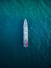 An overhead view of a large ship navigating through a vast expanse of deep blue ocean. The ship is centrally positioned, showcasing its streamlined design and various components visible on deck, including lifeboats and equipment.
