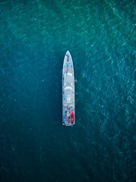 An overhead view of a large ship navigating through a vast expanse of deep blue ocean. The ship is centrally positioned, showcasing its streamlined design and various components visible on deck, including lifeboats and equipment.