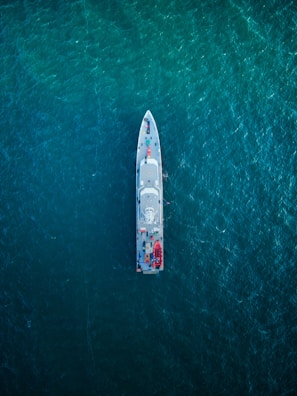 An overhead view of a large ship navigating through a vast expanse of deep blue ocean. The ship is centrally positioned, showcasing its streamlined design and various components visible on deck, including lifeboats and equipment.