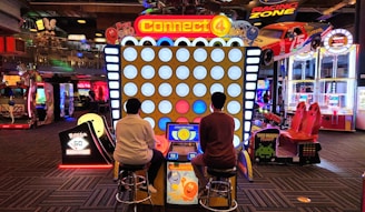 Two people are seated on stools playing an oversized version of the Connect 4 game in an arcade. Various other arcade games and machines are visible in the background, including racing games and a Monopoly-themed machine. The area is brightly lit with colorful lights.