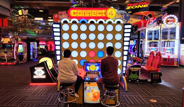Two people are seated on stools playing an oversized version of the Connect 4 game in an arcade. Various other arcade games and machines are visible in the background, including racing games and a Monopoly-themed machine. The area is brightly lit with colorful lights.