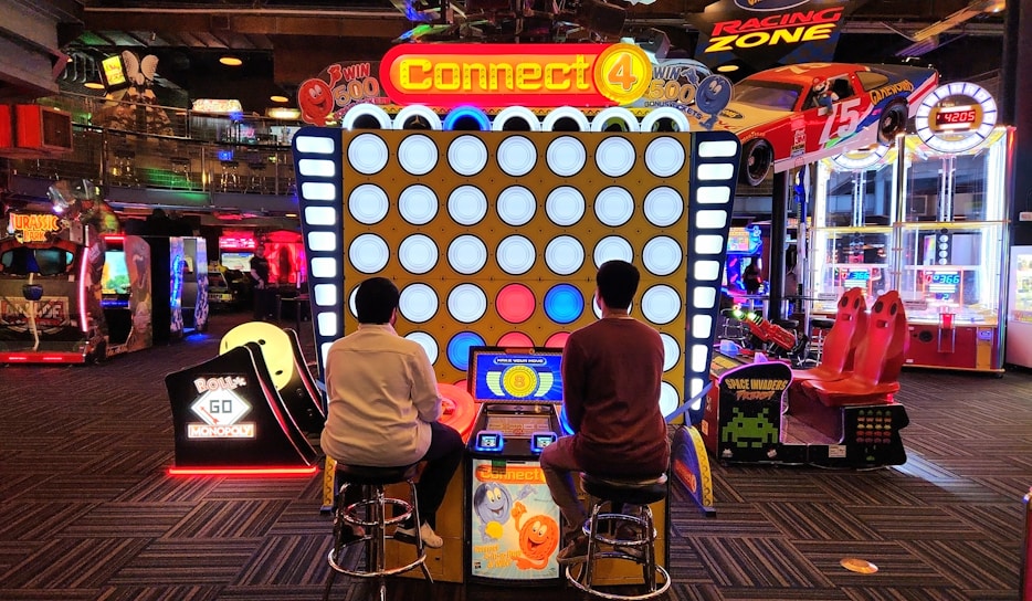Two people are seated on stools playing an oversized version of the Connect 4 game in an arcade. Various other arcade games and machines are visible in the background, including racing games and a Monopoly-themed machine. The area is brightly lit with colorful lights.