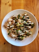 A white ceramic bowl contains roasted broccoli and cauliflower floret pieces drizzled with a creamy white sauce. The dish is garnished with fresh herbs and sprinkled with red seasoning. The bowl is placed on a wooden table with a varied wood grain pattern.