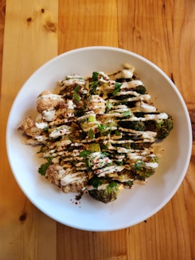 A white ceramic bowl contains roasted broccoli and cauliflower floret pieces drizzled with a creamy white sauce. The dish is garnished with fresh herbs and sprinkled with red seasoning. The bowl is placed on a wooden table with a varied wood grain pattern.