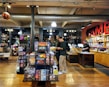 The image depicts the interior of a board game store with shelves filled with various games. Two customers stand near the counter, examining a game, while a cashier is present at the register. The warm lighting and wooden floors give the store a cozy atmosphere.