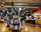 The image depicts the interior of a board game store with shelves filled with various games. Two customers stand near the counter, examining a game, while a cashier is present at the register. The warm lighting and wooden floors give the store a cozy atmosphere.