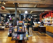 The image depicts the interior of a board game store with shelves filled with various games. Two customers stand near the counter, examining a game, while a cashier is present at the register. The warm lighting and wooden floors give the store a cozy atmosphere.