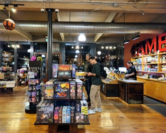 The image depicts the interior of a board game store with shelves filled with various games. Two customers stand near the counter, examining a game, while a cashier is present at the register. The warm lighting and wooden floors give the store a cozy atmosphere.