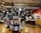 The image depicts the interior of a board game store with shelves filled with various games. Two customers stand near the counter, examining a game, while a cashier is present at the register. The warm lighting and wooden floors give the store a cozy atmosphere.
