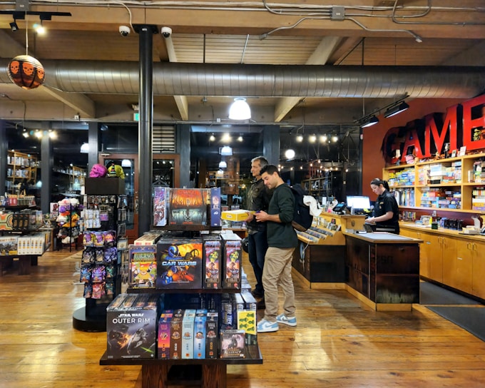 The image depicts the interior of a board game store with shelves filled with various games. Two customers stand near the counter, examining a game, while a cashier is present at the register. The warm lighting and wooden floors give the store a cozy atmosphere.